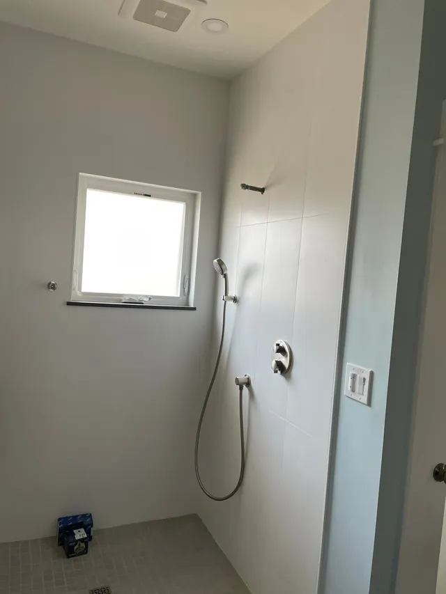 Small white tiled shower room with a window, handheld shower, and a soap dispenser on the wall.