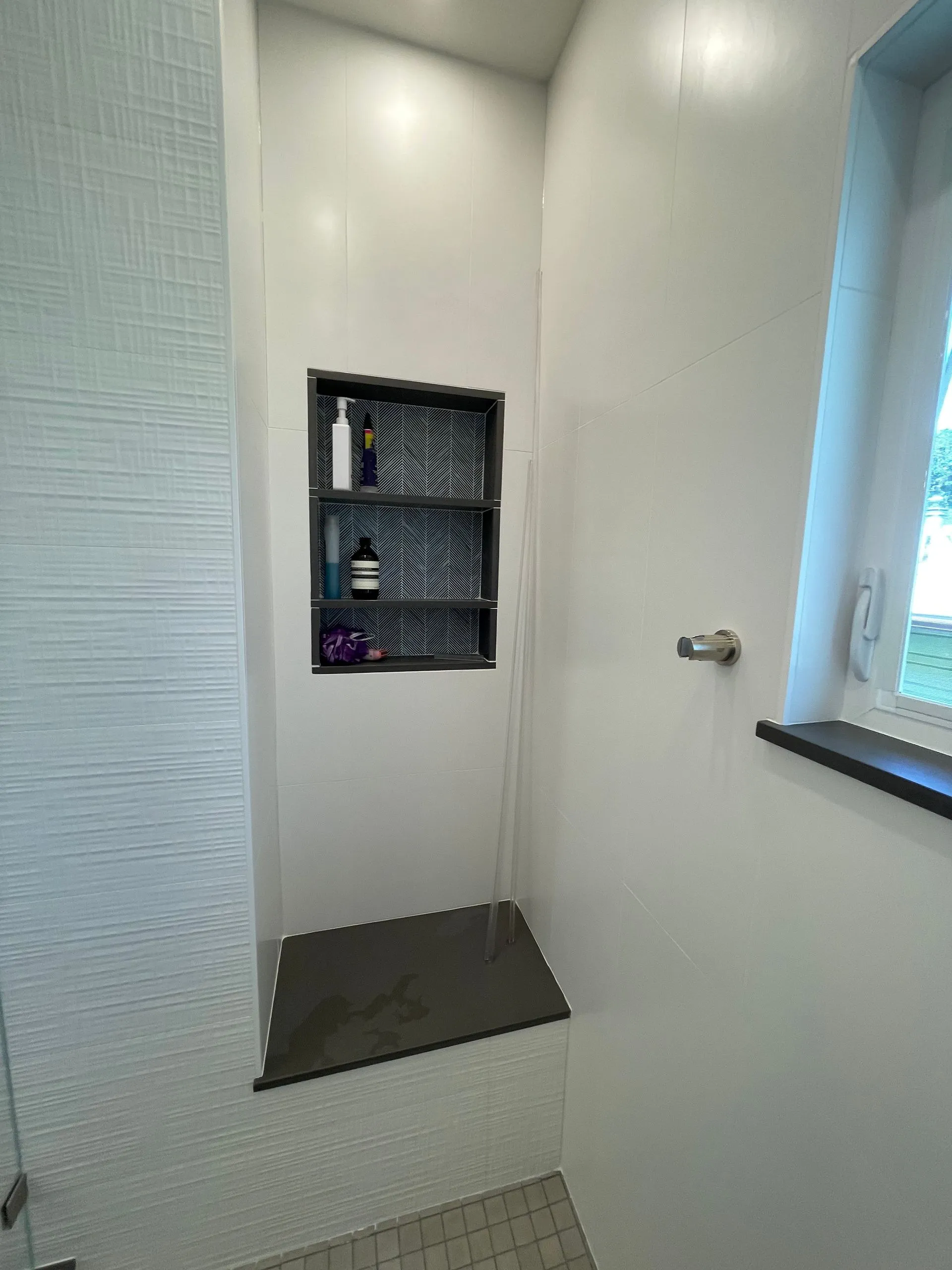 Built-in shower nook with gray tile shelf, dark floor ledge, and window light in a white bathroom