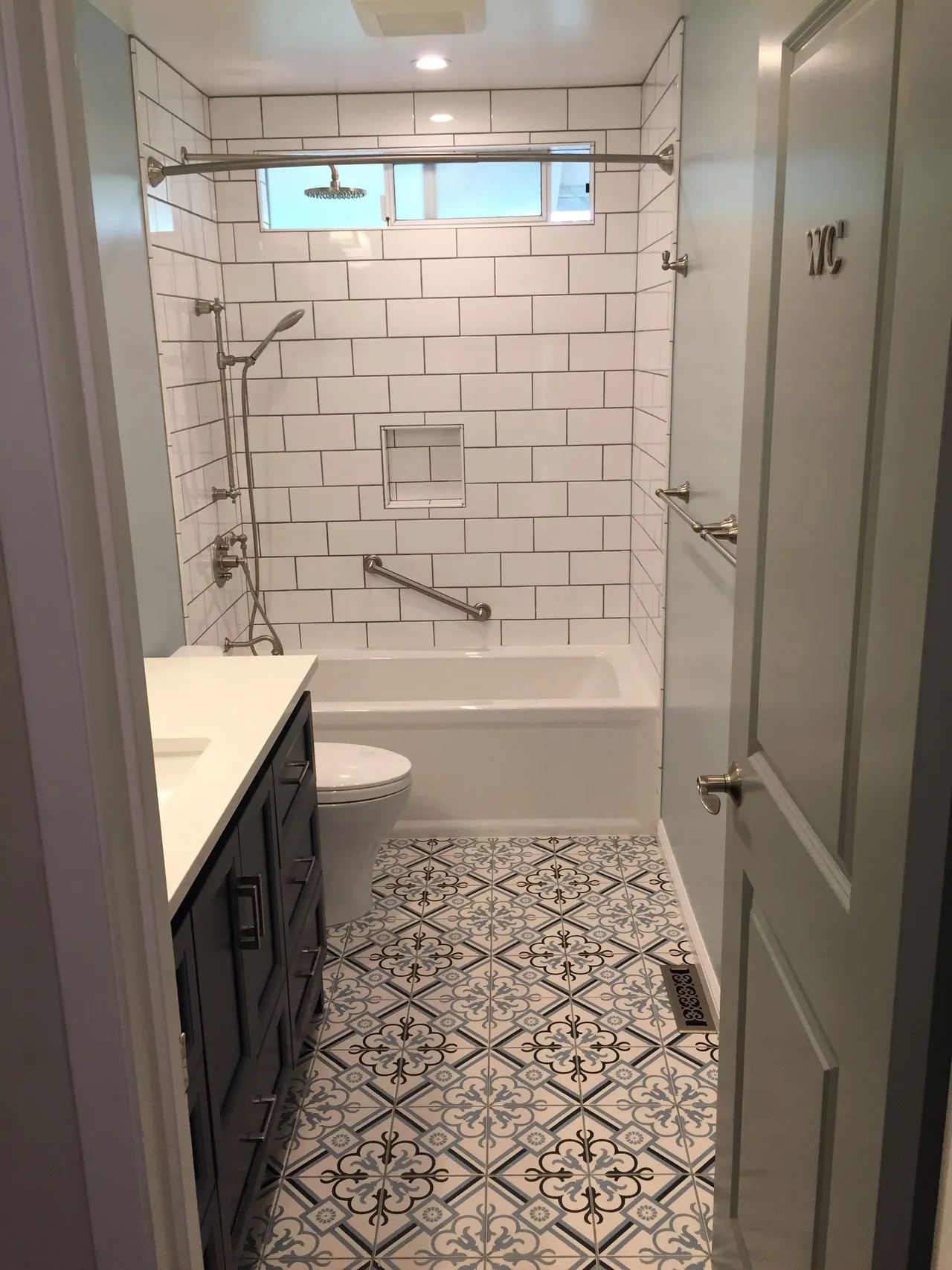 Narrow white bathroom with patterned tile floor, bathtub shower, toilet, vanity, and frosted glass door.