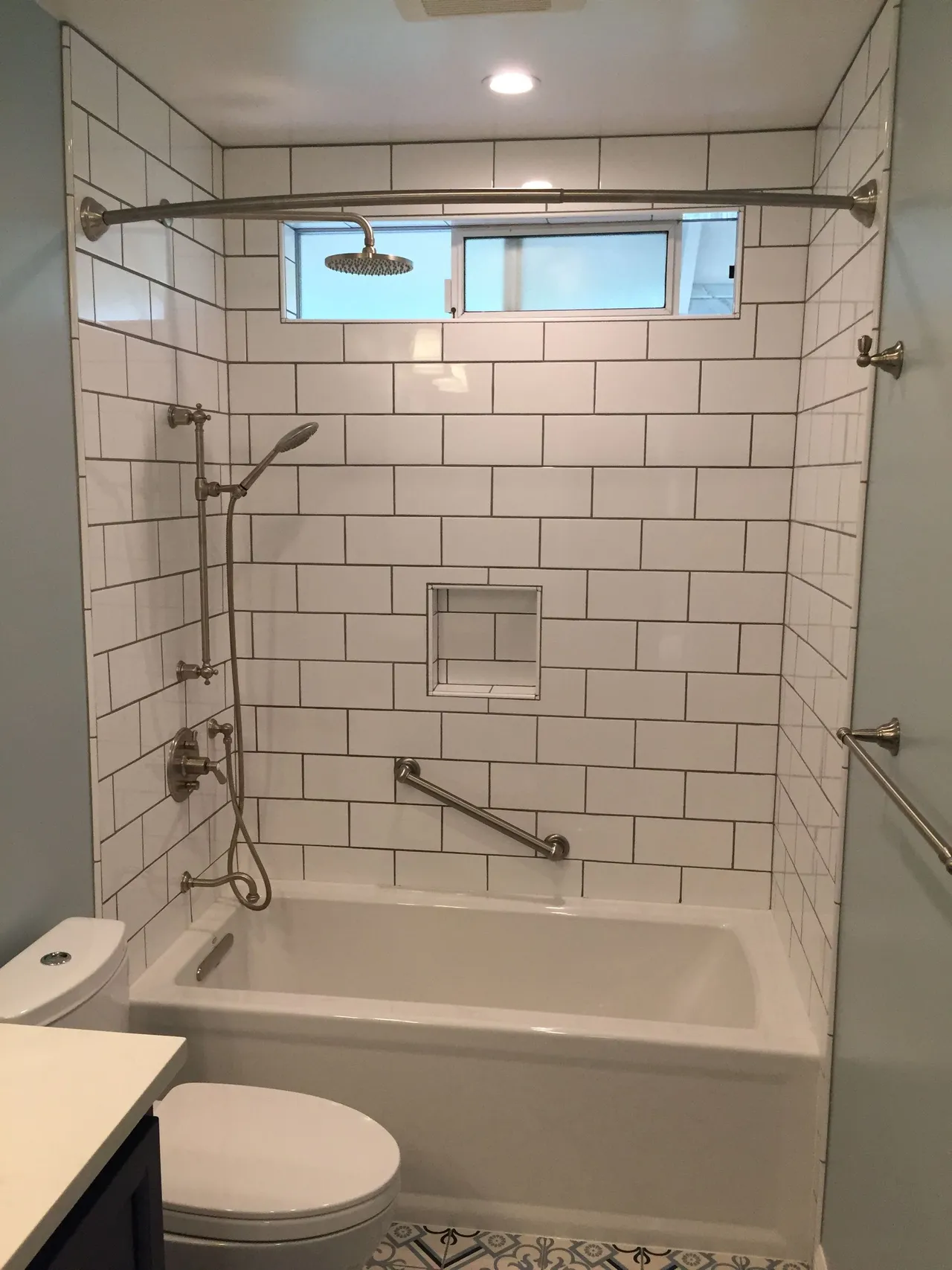 Accessible tiled bathroom with tub, grab bars, handheld shower, and toilet under a small window