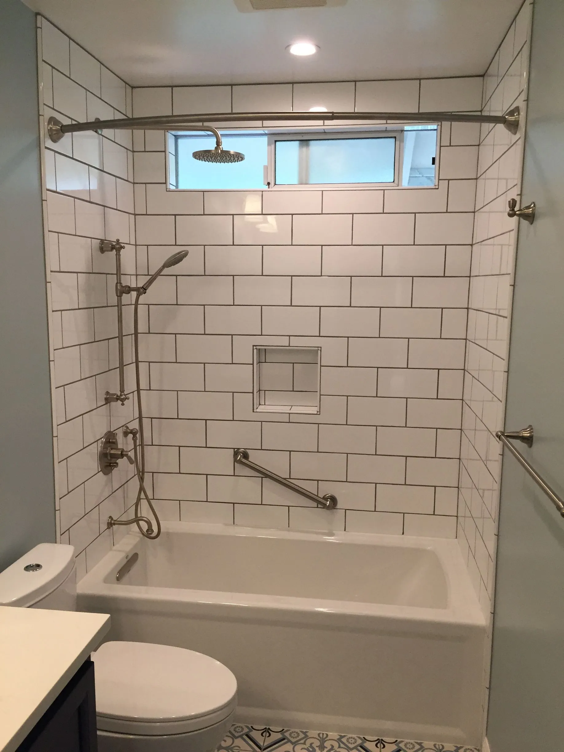Accessible tiled bathroom with tub, grab bars, handheld shower, and toilet under a small window