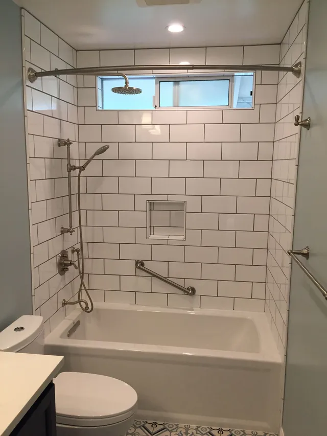Accessible tiled bathroom with tub, grab bars, handheld shower, and toilet under a small window
